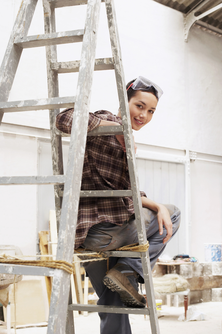 Portrait of happy young female painter sitting on ladder in work site